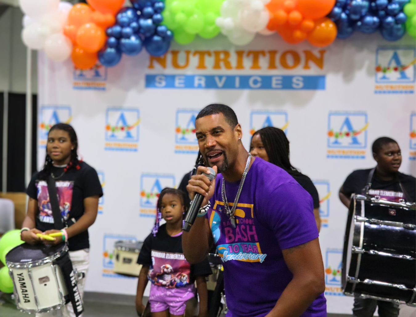 APS Nutrition Services representative speaking on stage in front of students and drummers at the Back to School Bash